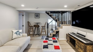 A modern living room with a sofa, a coffee table with cushions, a piano, a TV, and a staircase leading to another room.