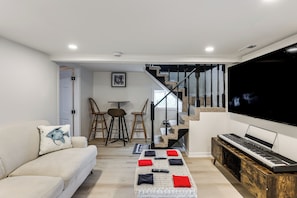 Una sala moderna con sofá, una mesa de centro con cojines, un piano, un televisor y una escalera que lleva a otra habitación.