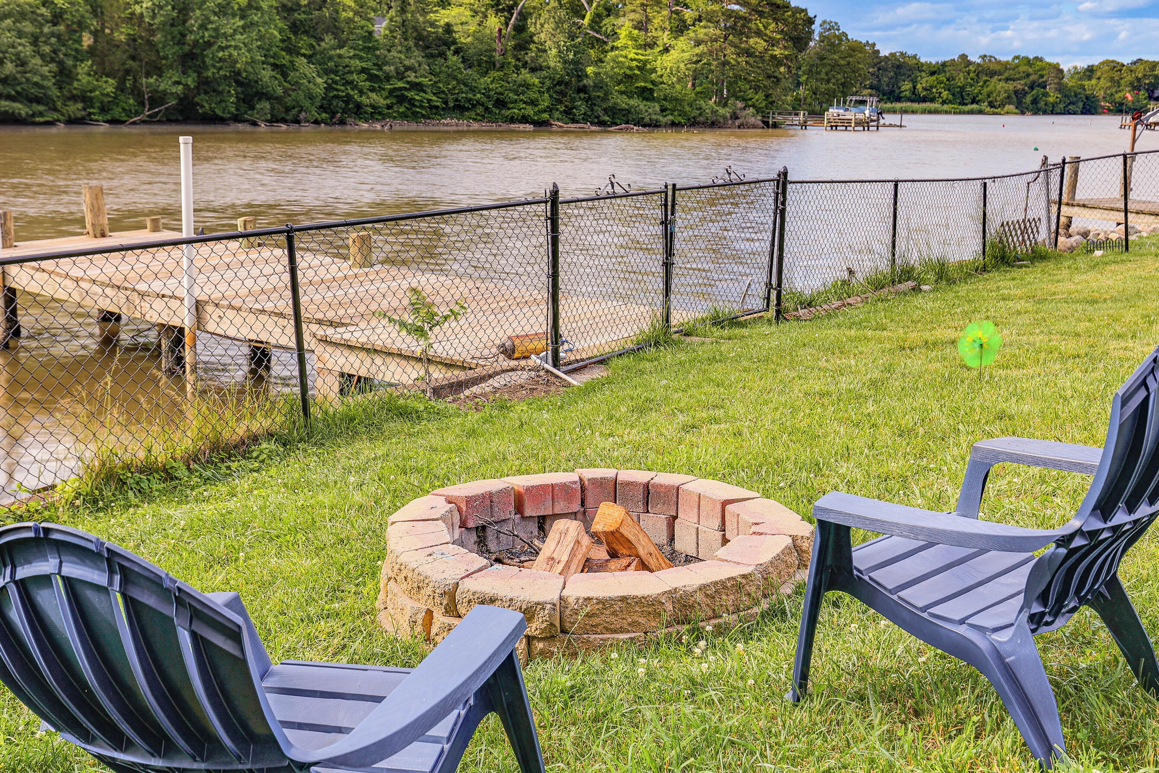 A riverside deck with a fire pit and Adirondack chairs, surrounded by a chain-link fence and a dock.