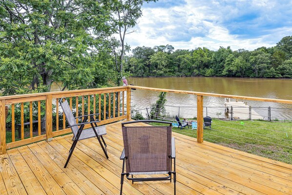 A wooden deck with a chair and a table, overlooking a lake with a grassy area and a fence.