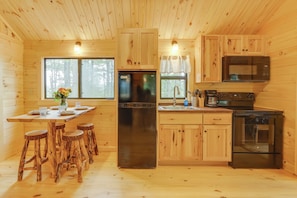 A wooden cabin kitchen with a central island, stainless steel appliances, and a large window with a view of trees. - Stunning Views! 3-cabin Retreat on 400-acre Farm (Middlebury Center)