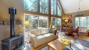 A spacious living room with a large window, a fireplace, a sofa, a coffee table, and a dining area. - Projector & Deck! Family Cabin, 4 Mi to Lake Davis (Portola)