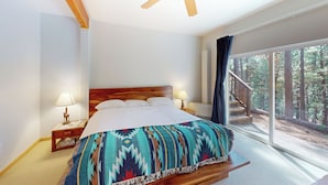 A bedroom with a large bed, wooden headboard, two bedside tables with lamps, a ceiling fan, and a large window with a forest view. - Projector & Deck! Family Cabin, 4 Mi to Lake Davis (Portola)