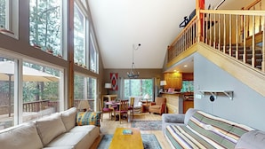 A spacious living room with a sofa, a coffee table, and a striped couch. There is a dining area with chairs and a table, and a kitchen area with wooden cabinets. - Projector & Deck! Family Cabin, 4 Mi to Lake Davis (Portola)