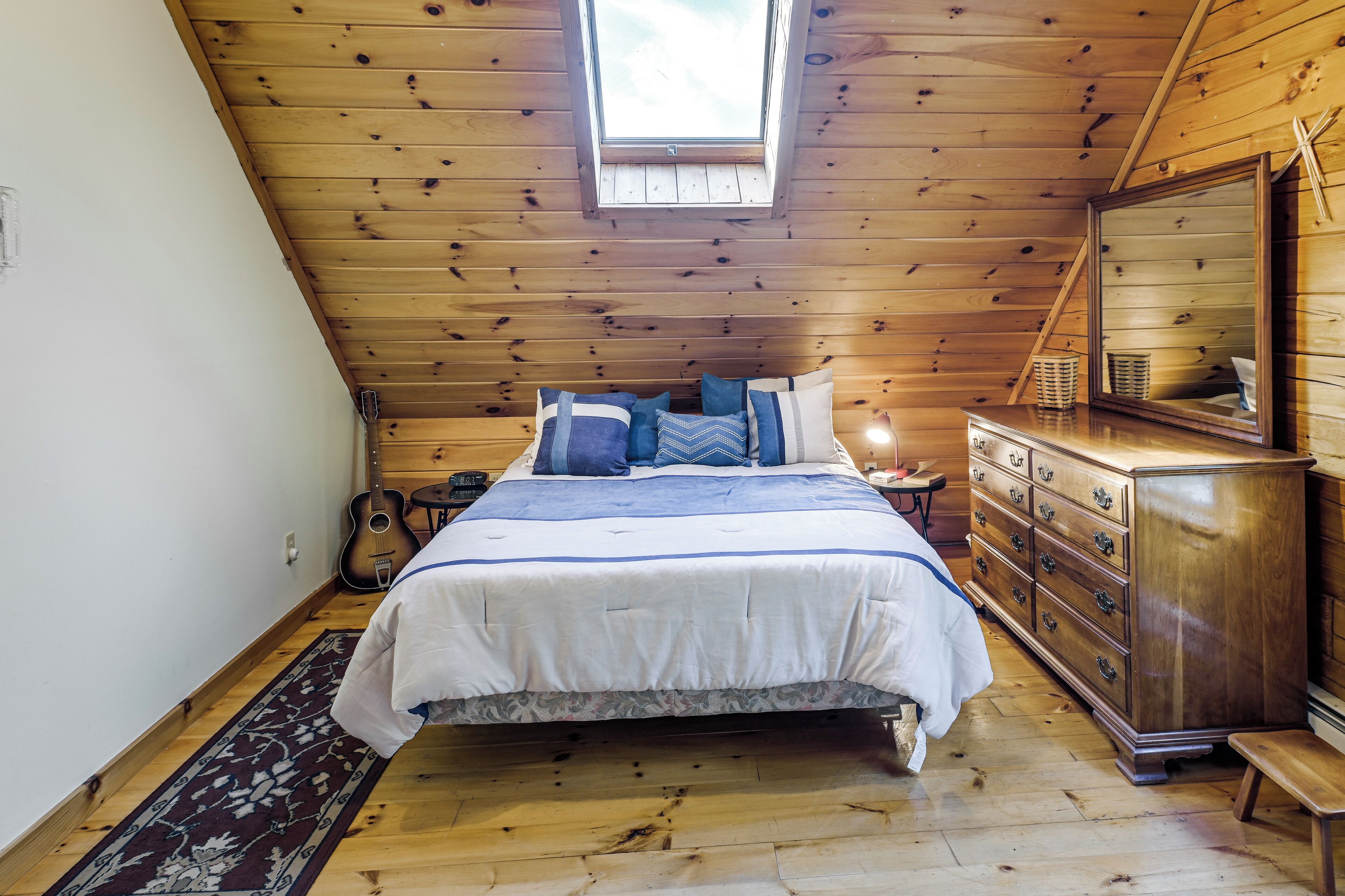 A bedroom with a wooden ceiling, a skylight, a bed with blue and white bedding, a dresser, and a guitar.