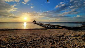 Ein Strand mit einer hölzernen Pier, die sich in ruhiges Wasser erstreckt, eine kleine Hütte und ein klarer Himmel.