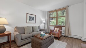 Een woonkamer met een bank, poef, salontafel, fauteuil, lamp, raam en kunstwerk.