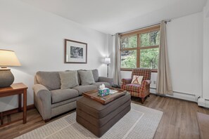 A living room with a sofa, ottoman, coffee table, chair, lamp, window, and artwork. - Zephyr Mountain Lodge Unit 1407 Private deck ski-in/ski-out on site amenities (Winter Park)