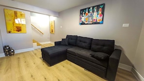 A living room with a sectional sofa, a small ottoman, a staircase, and a wall with a bottle art piece.