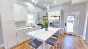 A modern kitchen with white cabinets, a large island with stools, a stainless steel refrigerator, and a window providing natural light.