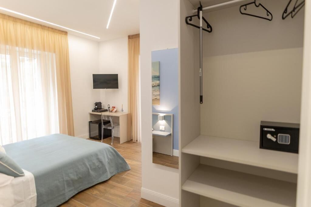 A hotel room with a bed, a desk with a computer, a TV, and a wardrobe with hangers.