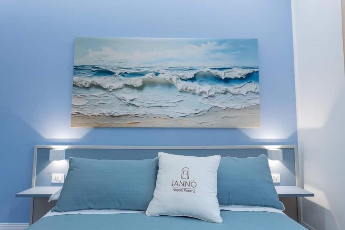 A bedroom with a bed, two pillows, a wall-mounted painting of waves, and a bedside table with a lamp.