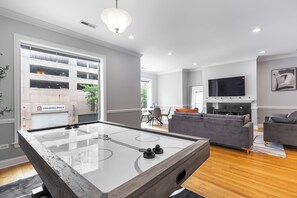 Ein modernes Wohnzimmer mit einem Kicker, grauen Sofas, einem Flachbildfernseher und einem Essbereich im Hintergrund.