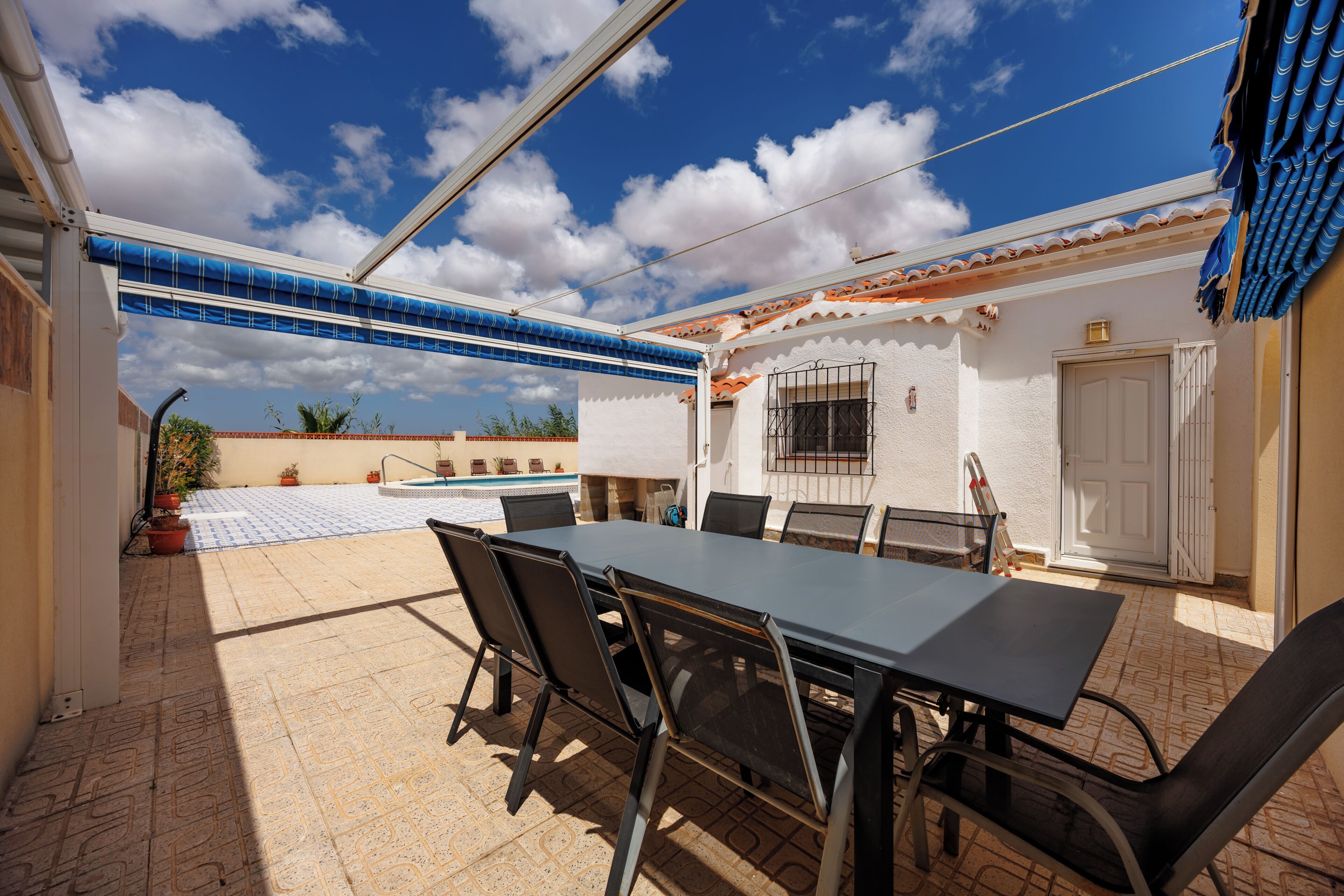 Eine Terrassenausstattung im Freien mit Tisch und Stühlen, ein Schwimmbecken und ein weißes Gebäude mit einem gefliesen Dach.
