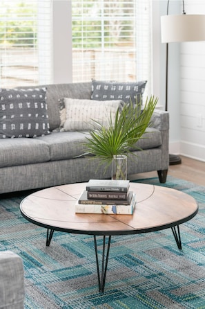 Ein Wohnzimmer mit grauem Sofa, einem runden Couchtisch mit Büchern und einer Pflanze sowie einem gemusterten Teppich.