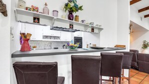 A kitchen with white cabinets, a central island with bar stools, and decorative items on shelves. - Little Flower House | Heated Cowboy Pool, Fire Pit & Minutes to the Square (Wimberley)