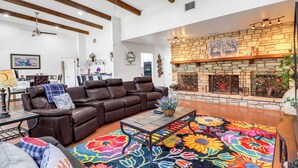 A living room with a brown leather sofa, a colorful floral rug, a stone fireplace, and a ceiling fan. - Little Flower House | Heated Cowboy Pool, Fire Pit & Minutes to the Square (Wimberley)