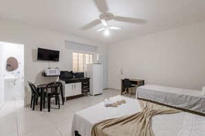 Quarto de hotel com cama, escrivaninha com computador, televisão, ventoinha no teto e janela.