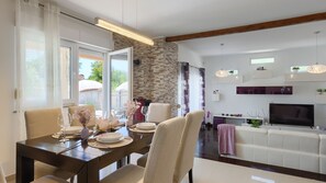 A dining area with a table set for four, a kitchen area with a TV, and a stone wall. - Villa Elssa by Villas Guide (Ližnjan)