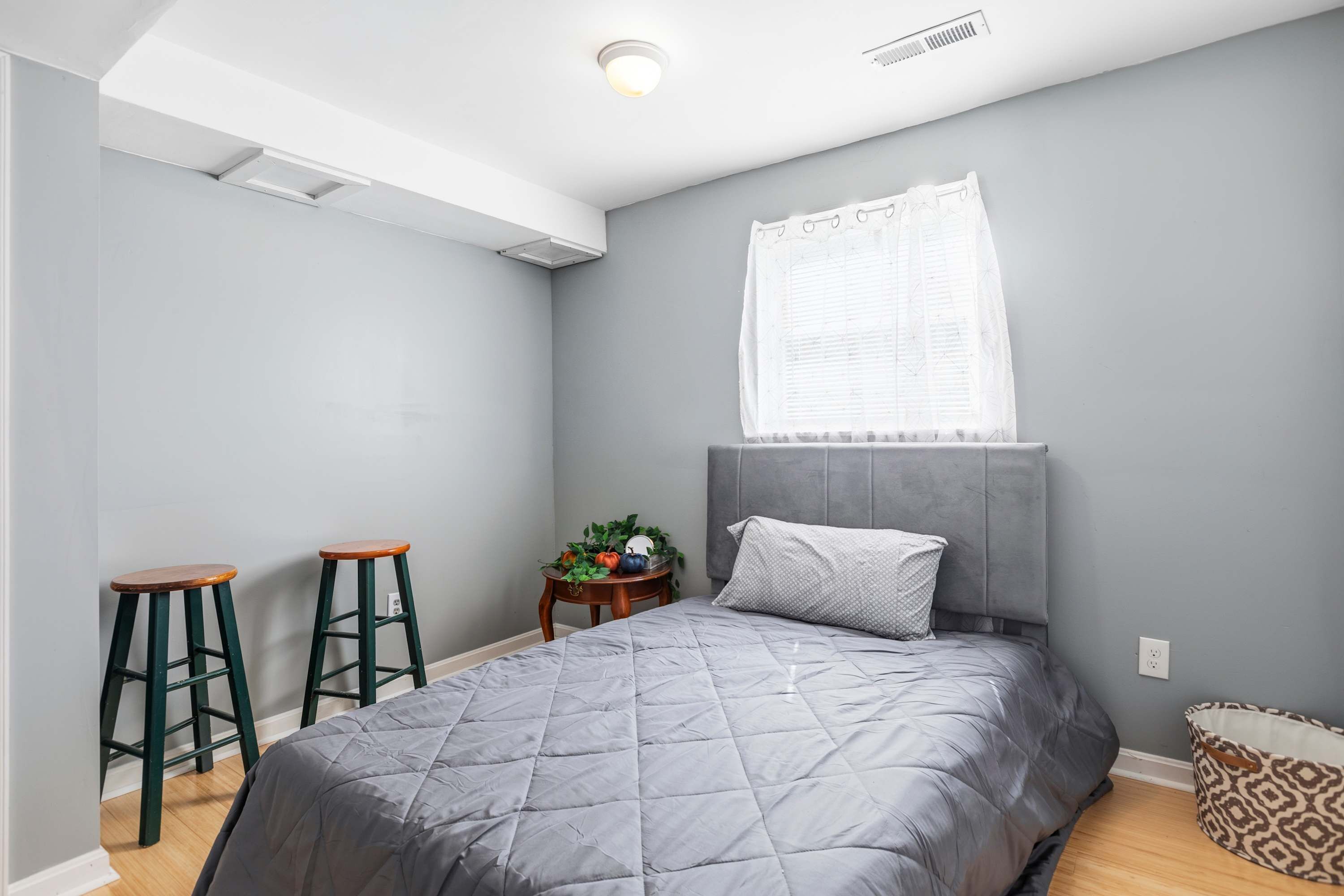 A bedroom with a grey bed, two wooden stools, a small table with plants, a grey headboard, and a window with white curtains.