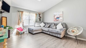 Una sala de estar moderna con un sofá seccional, un sillón blanco, un televisor de pantalla plana y un ventanal con cortinas.