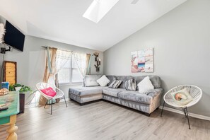 A modern living room with a sectional sofa, a white chair, a flat-screen TV, and a large window with curtains. - Lusby 5BD Family Home With Private Beach Access (Lusby)