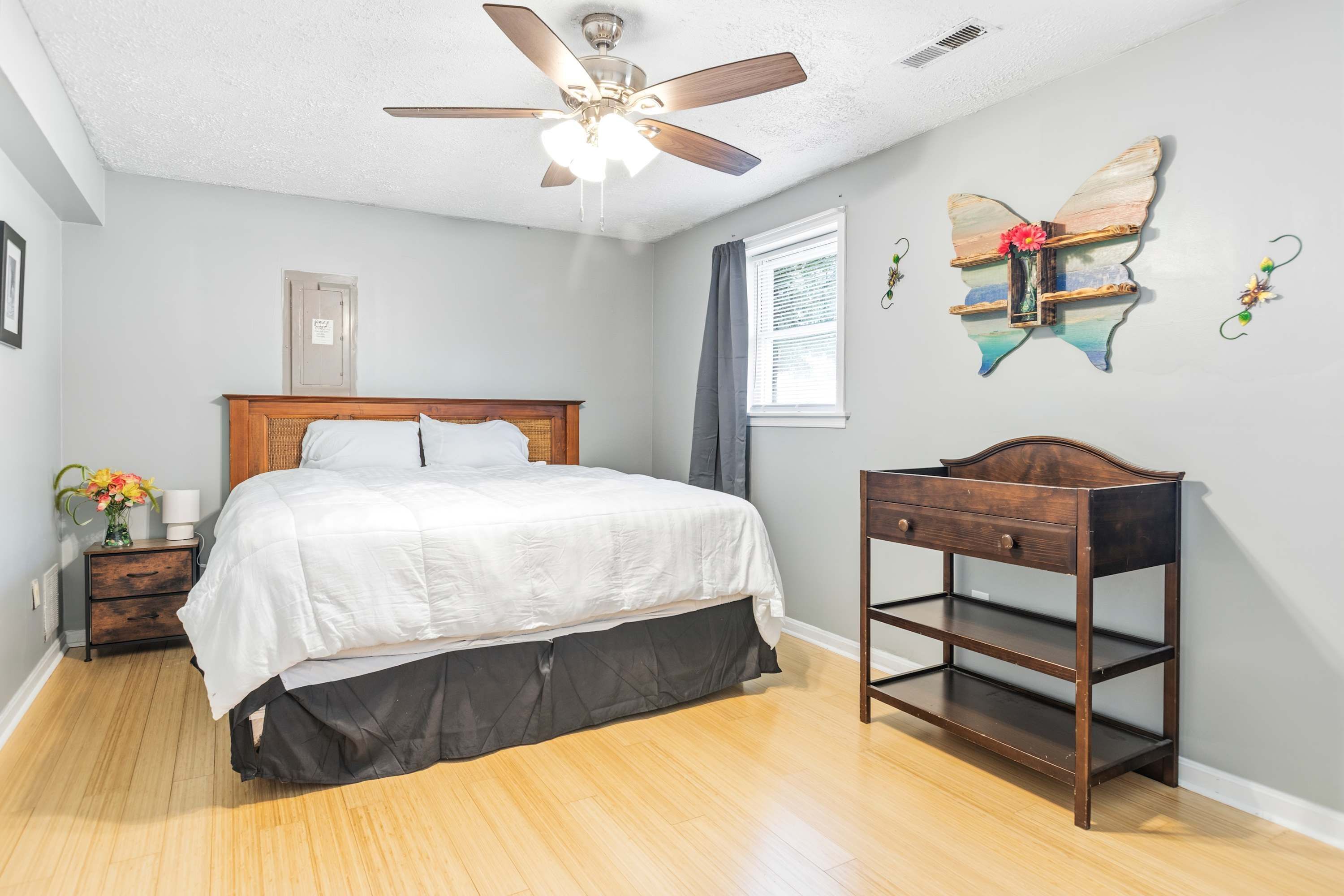 A bedroom with a bed, a nightstand, a dresser, a ceiling fan, a window with curtains, and a wall-mounted butterfly decoration.