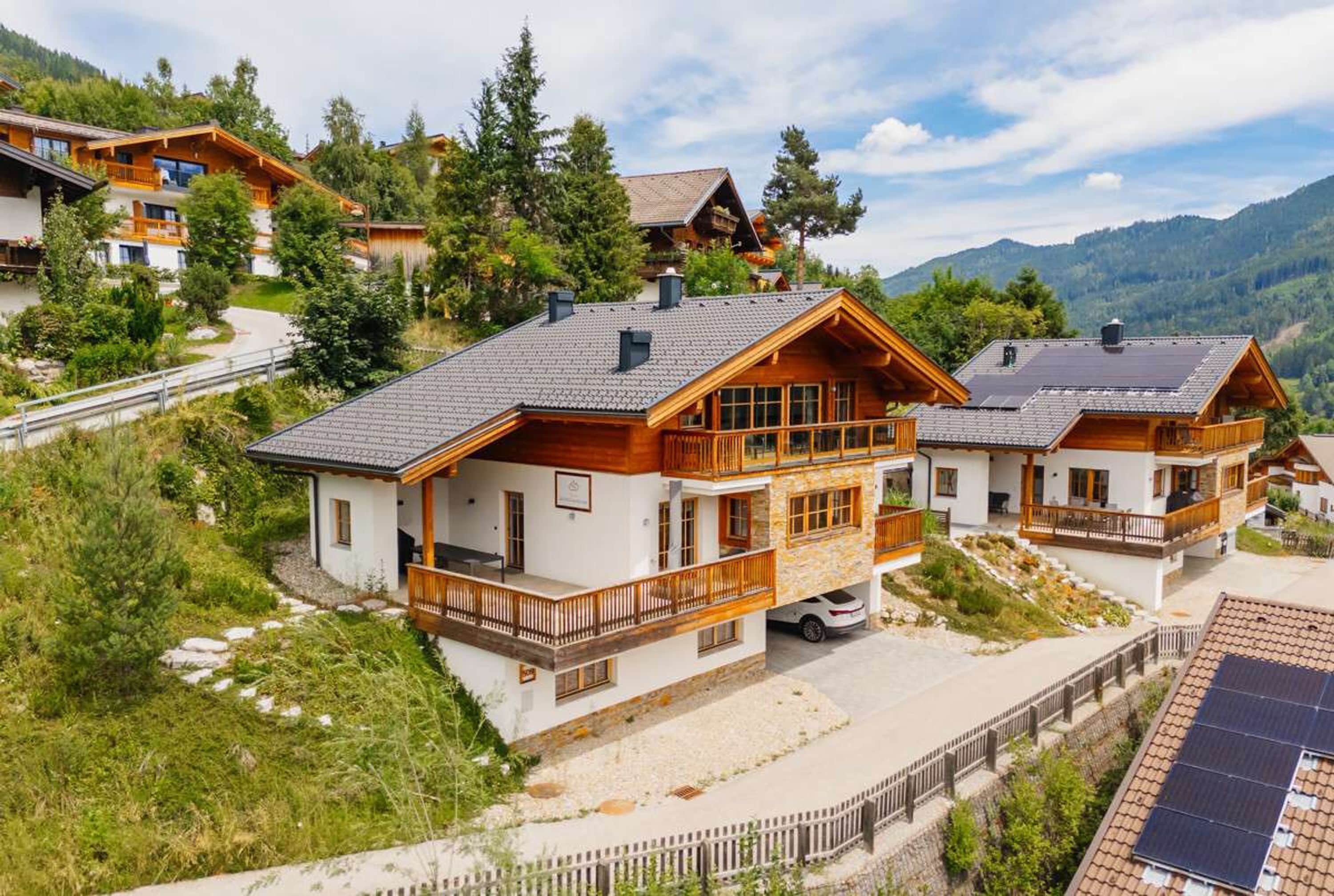 A chalet-style building with a balcony, a car parked in front, and a mountainous backdrop.