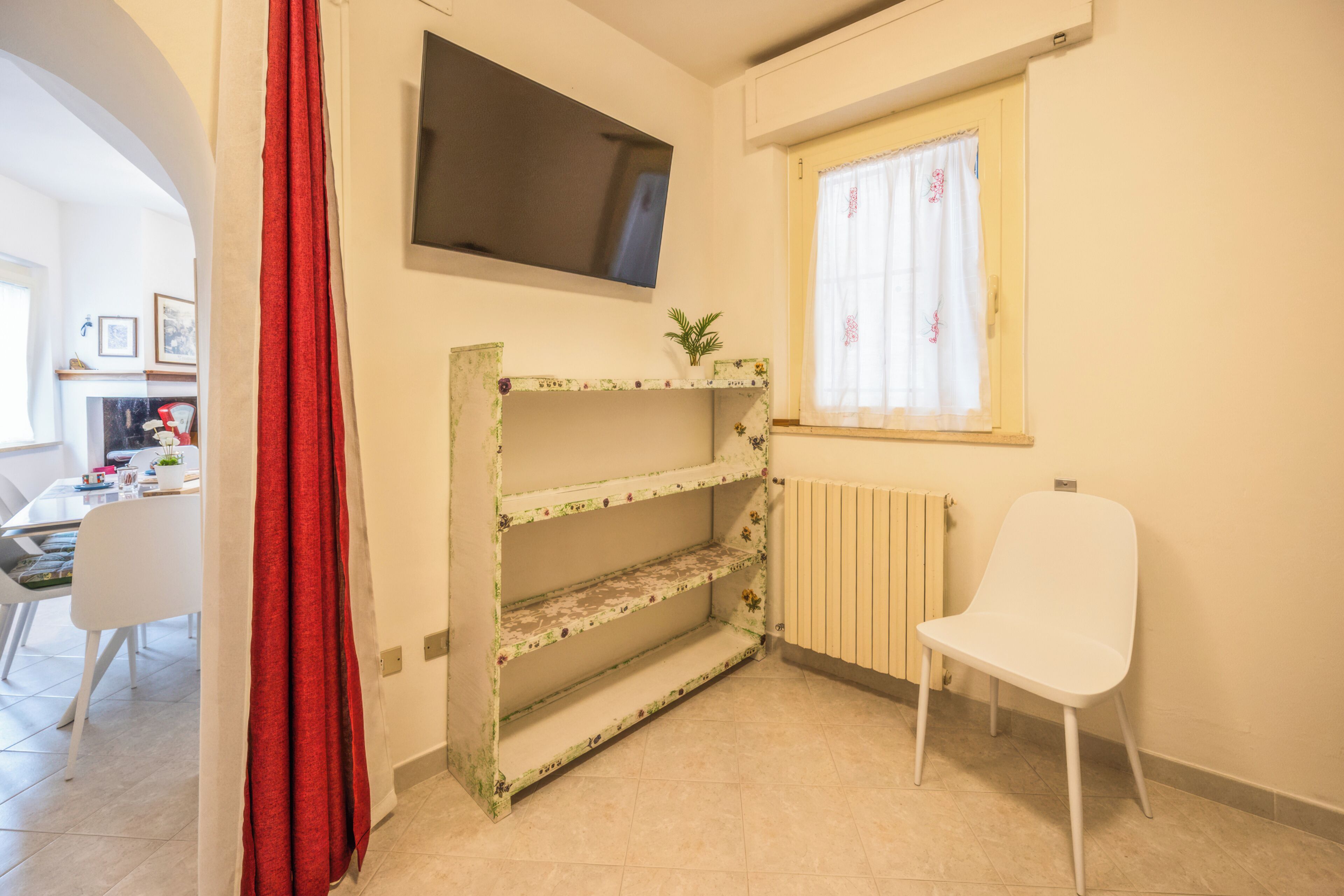 A room with a TV mounted on the wall, a radiator, a window with curtains, a white chair, and a bookshelf with floral patterns.