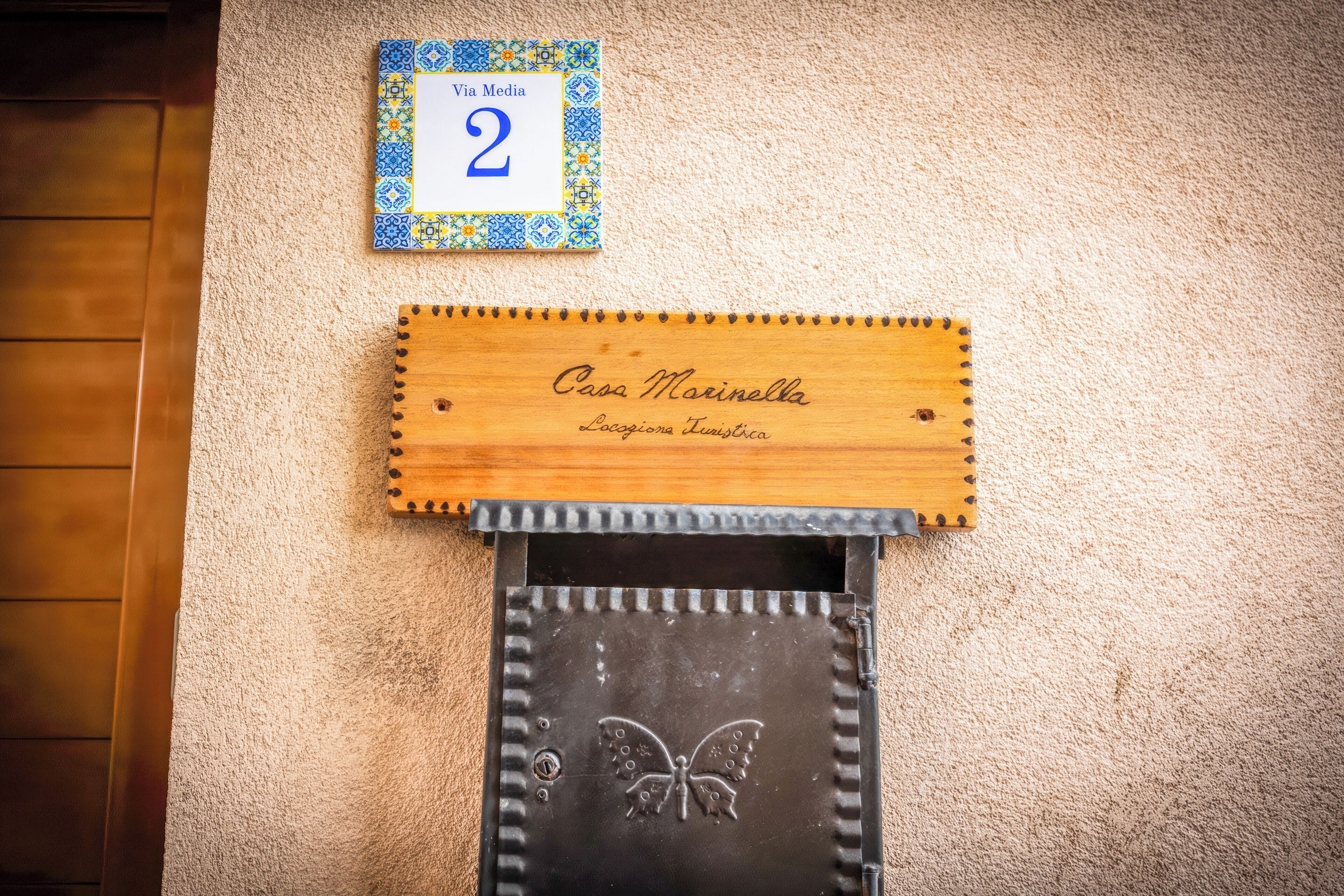 Una placa de madera con el nombre 'Casa Marinella' y 'Lazionario Turistico' grabado, un cartel numerado 'Via Media 2', y una pared negra con un diseño de mariposa.