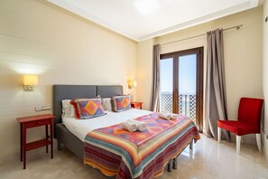 Ein Hotelzimmer mit einem Bett, einem roten Sessel, einem Nachttisch und einem Balkon mit Vorhängen.
