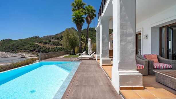 Ein Pool mit Holzterrasse, weißen Säulen und einer Korbsesselgarnitur mit Kissen.