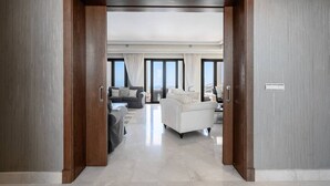 Ein modernes Hotelzimmer mit Holztür, einem Wohnbereich mit Sofa und Sesseln, einem Essbereich und großen Fenstern.