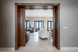 Ein modernes Hotelzimmer mit Holztür, einem Wohnbereich mit Sofa und Sesseln, einem Essbereich und großen Fenstern.