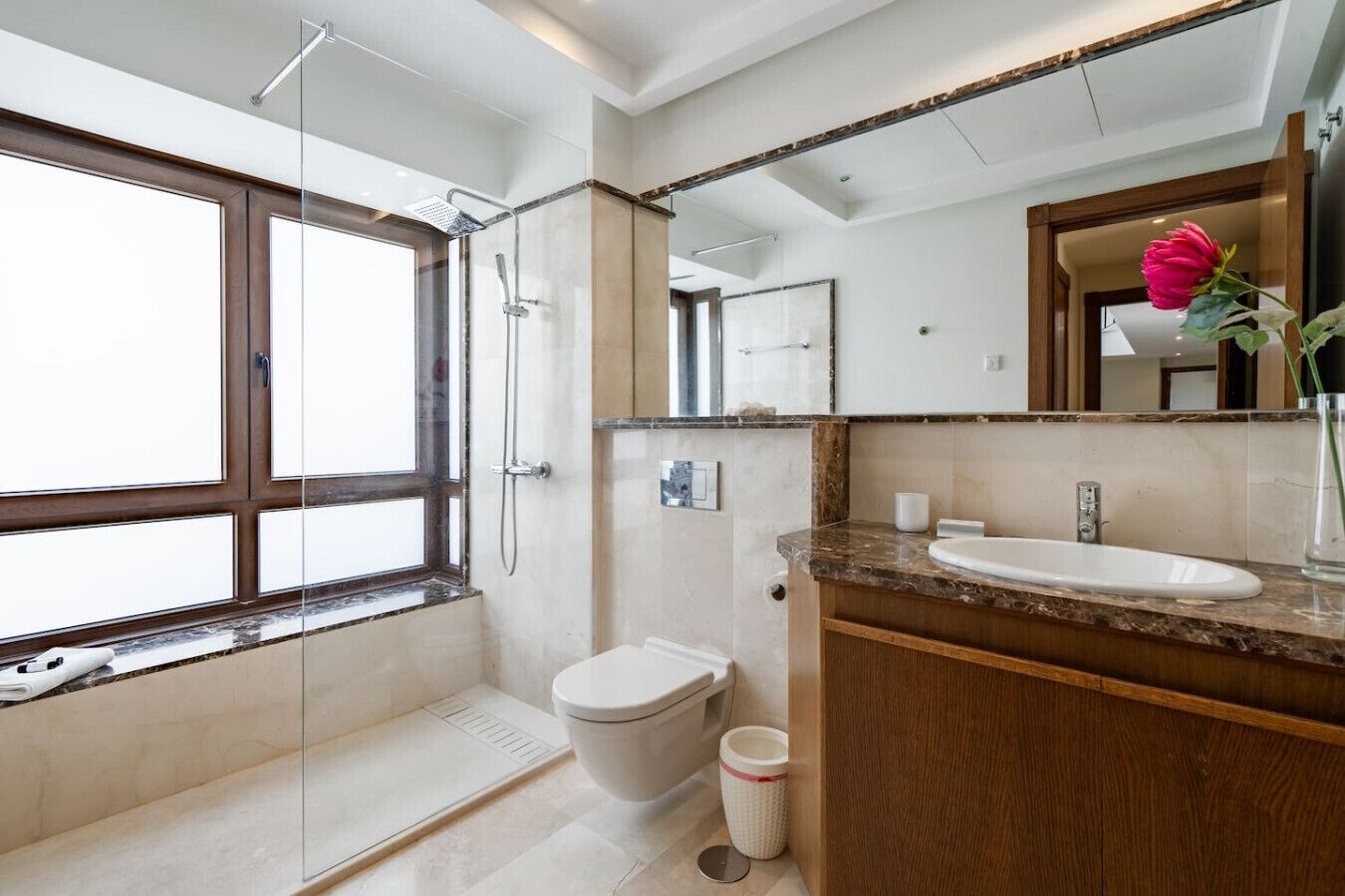A modern bathroom with a glass shower enclosure, a toilet, a sink with a marble countertop, and a large mirror.