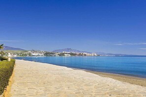 A paved walkway along a coastal area with a clear view of the sea and distant buildings. - ESTEPONA SUNSETS - BRAND-NEW MODERN 3-BEDROOM APARTMENT (Estepona)