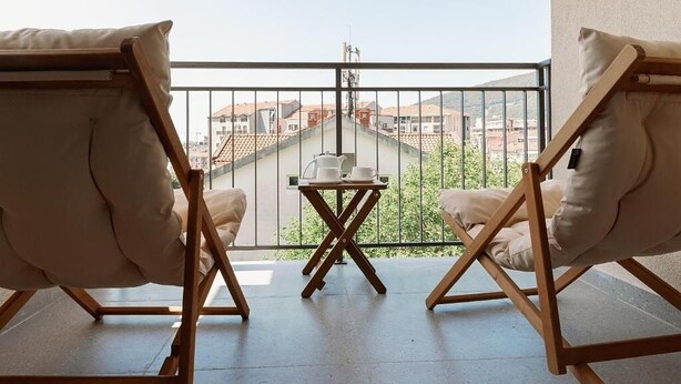 Ein Balkon mit zwei gepolsterten Stühlen, einem kleinen Tisch mit Teekanne und Tassen sowie Blick auf Gebäude.