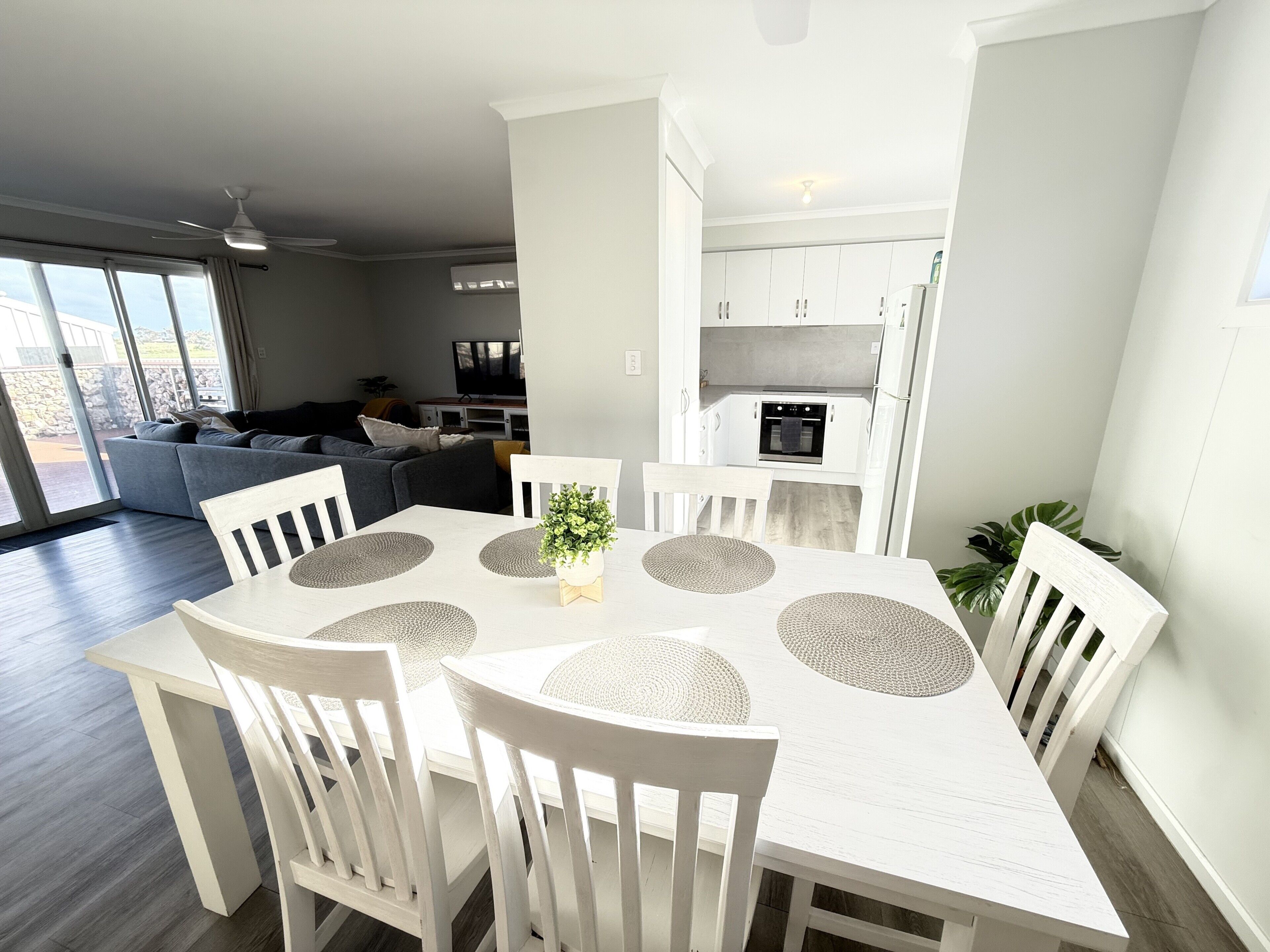 Un área de comedor moderna con una mesa y sillas blancas, una cocina al fondo y un sofá gris en la sala de estar.
