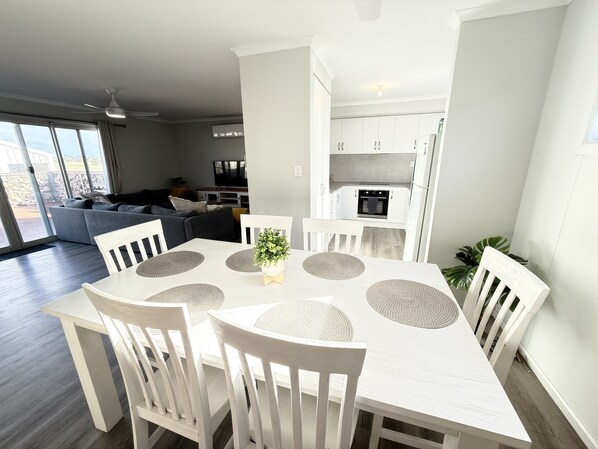 A modern dining area with a white table and chairs, a kitchen in the background, and a grey sofa in the living room.