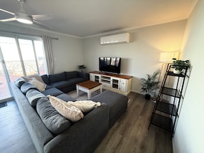 A living room with a grey sectional sofa, a wooden coffee table, a TV on a white cabinet, a ceiling fan, and a potted plant.