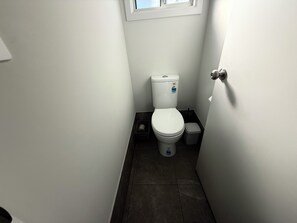 Une petite salle de bain propre, avec des toilettes blanches, une poubelle et une fenêtre située au-dessus des toilettes.