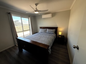 A bedroom with a bed, a ceiling fan, an air conditioning unit, a nightstand with a lamp, and a window with curtains.