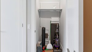 A narrow hallway with a white door, a shelf with a box, a vacuum cleaner, a broom, and a trash bag.