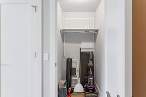 A narrow hallway with a white door, a shelf with a box, a vacuum cleaner, a broom, and a trash bag.