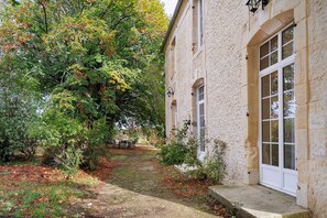 Ein Steinhaus mit großem Fenster und einer Tür, umgeben von Bäumen und einem Gartenweg.