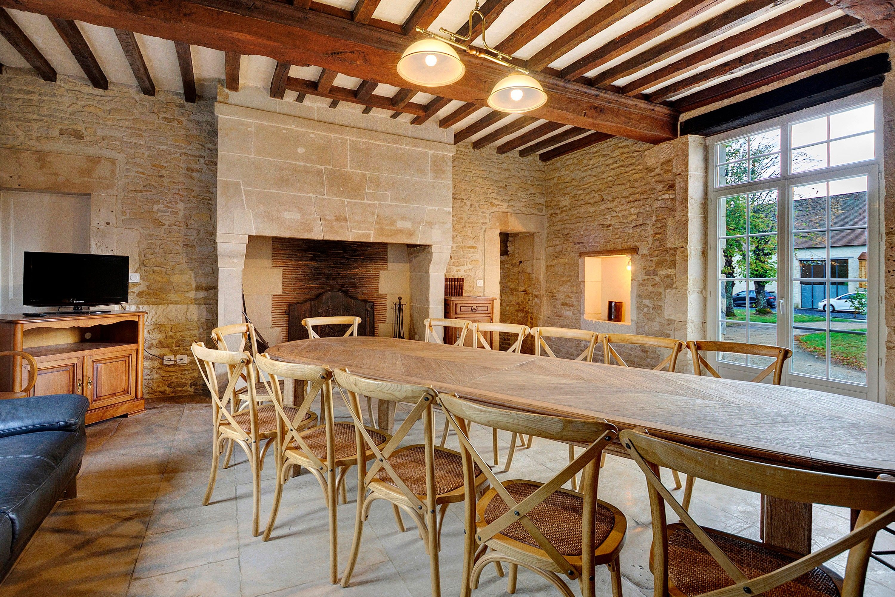 A spacious dining area with a long wooden table and wicker chairs, a stone fireplace, and a television on a wooden cabinet.