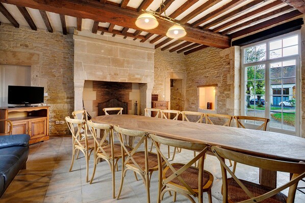A spacious dining area with a long wooden table and wicker chairs, a stone fireplace, and a television on a wooden cabinet.