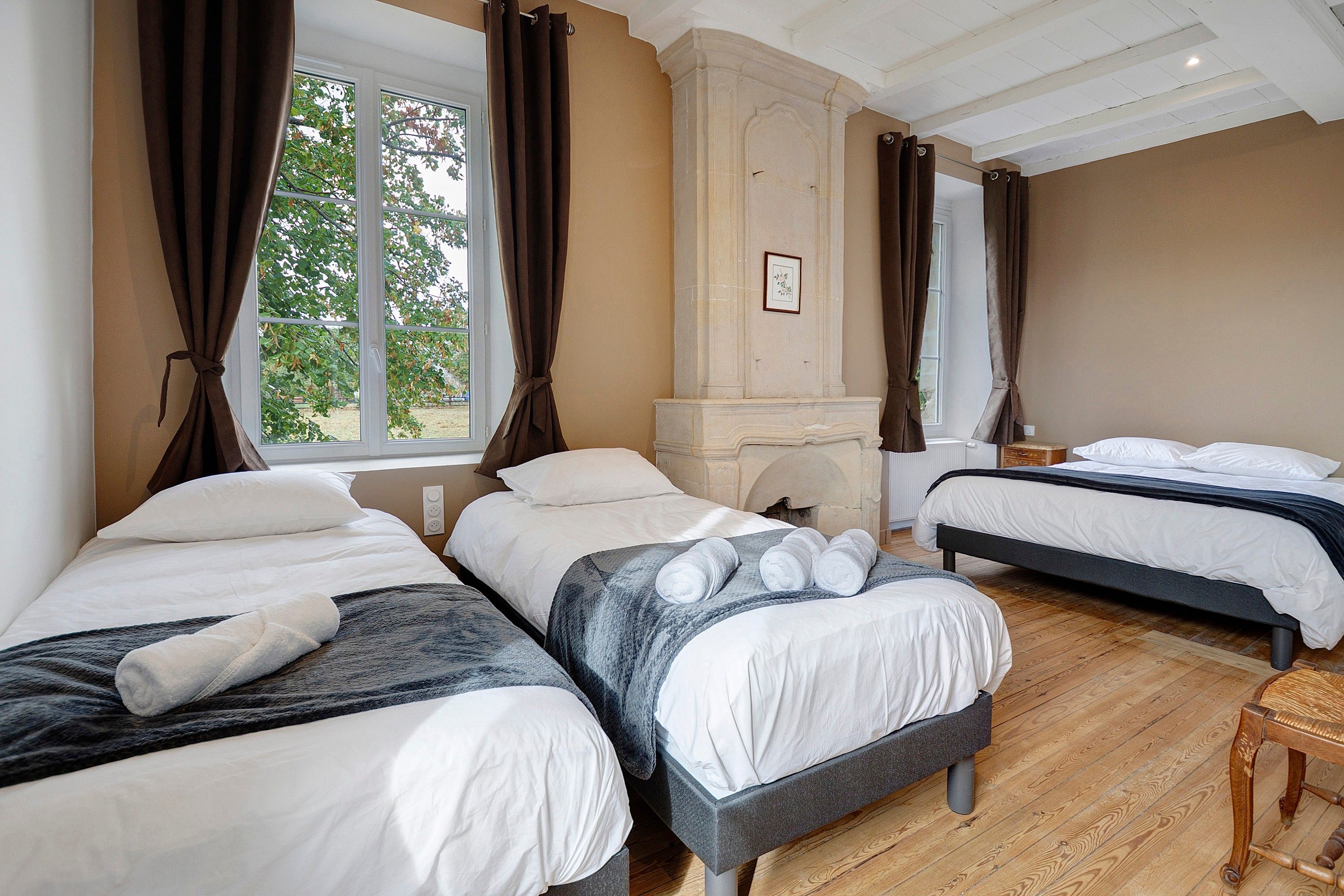A hotel room with three beds, a fireplace, and large windows with curtains.