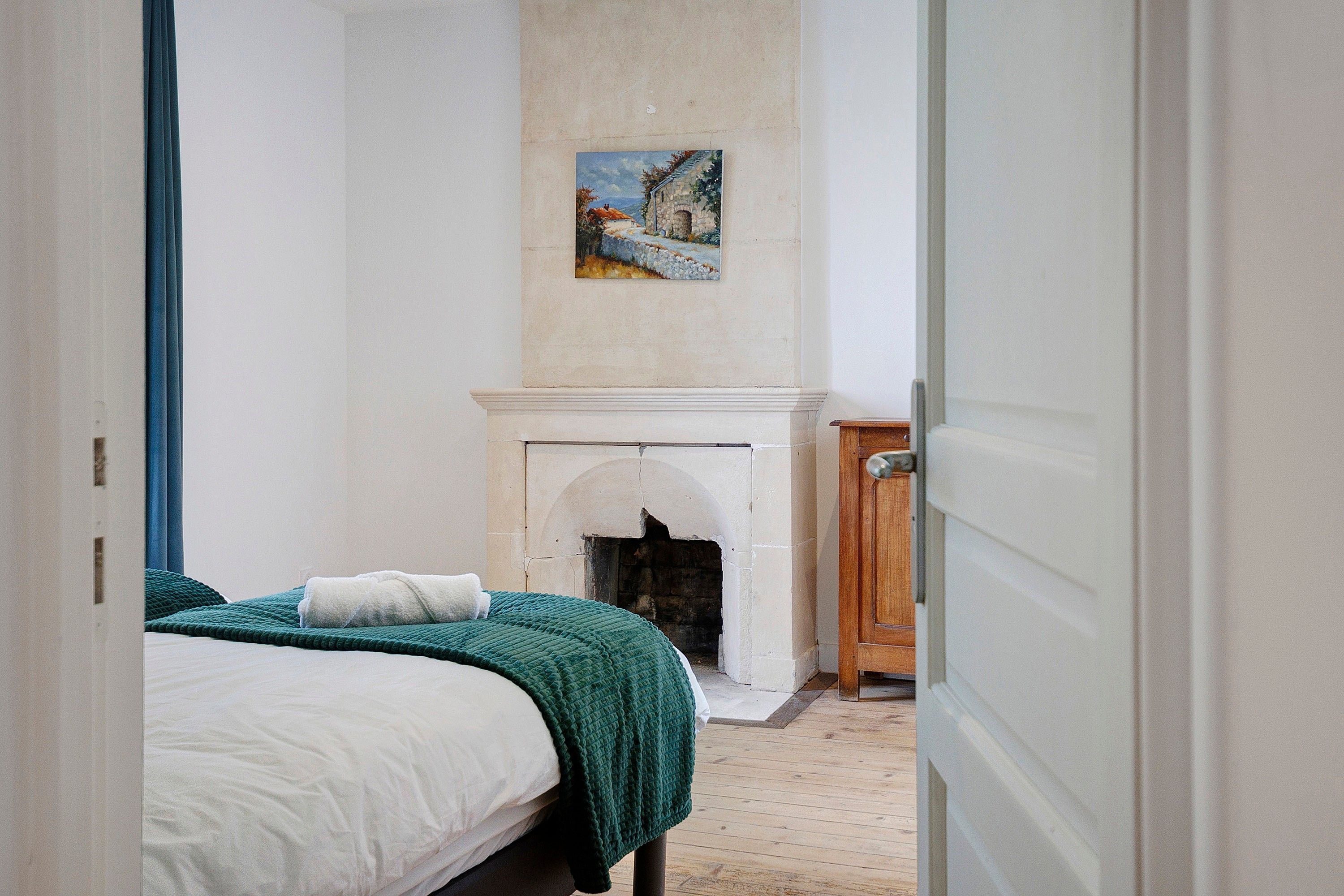 Een smalle gang die leidt naar een slaapkamer met een bed, een open haard en een schilderij aan de muur.