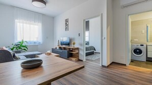 A modern living room with a sofa, TV, and dining table. There's a laundry area with a washing machine and dryer.